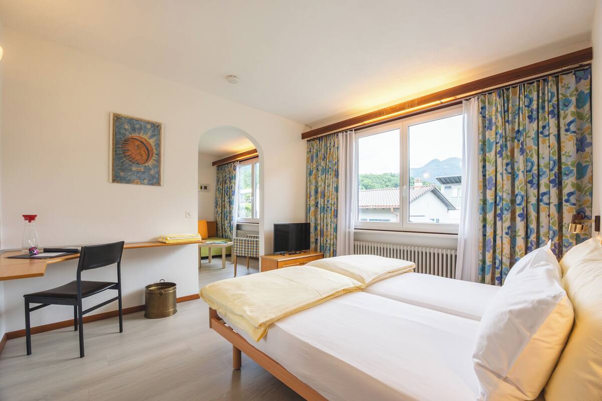 Quarto de hotel com cama, escrivaninha, cadeira, televisão e janela com cortinas.