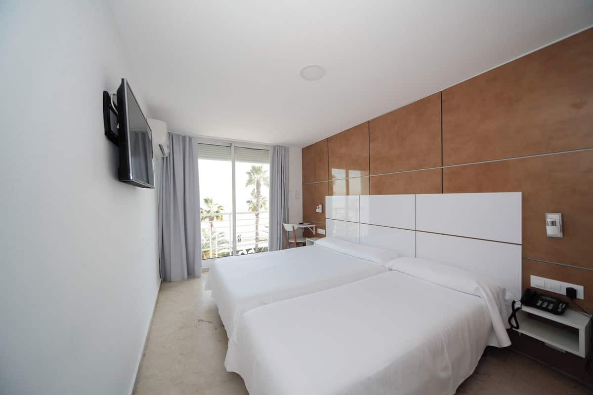 A hotel room with two beds, a TV mounted on the wall, a balcony with a view of palm trees, and a wooden accent wall.