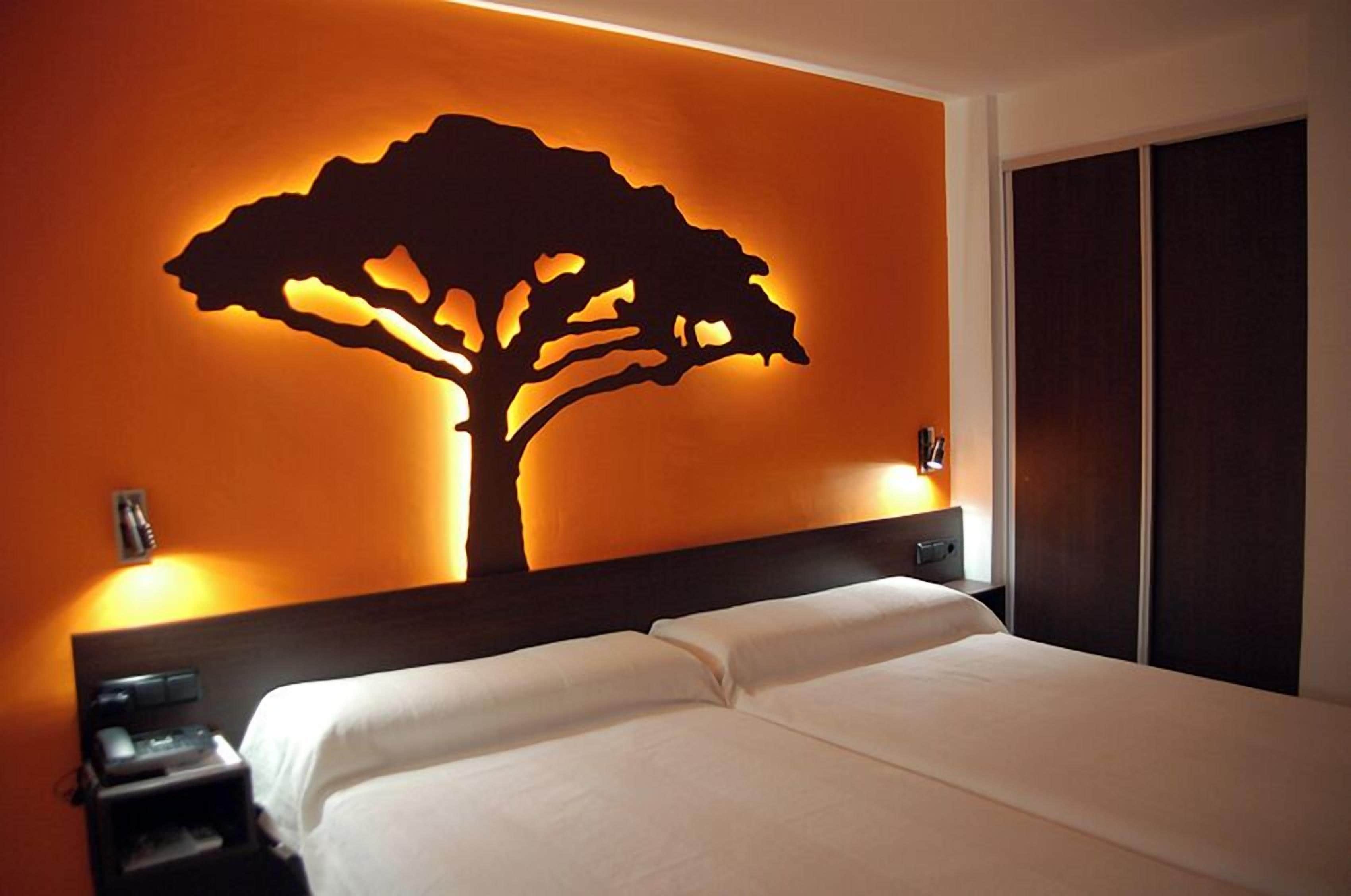 A hotel room with a bed, a nightstand, a wall-mounted tree silhouette, and a wardrobe.