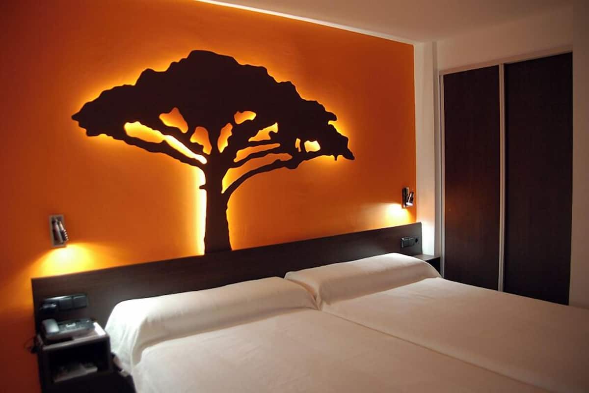 A hotel room with a bed, a nightstand, a wall-mounted tree silhouette, and a wardrobe.