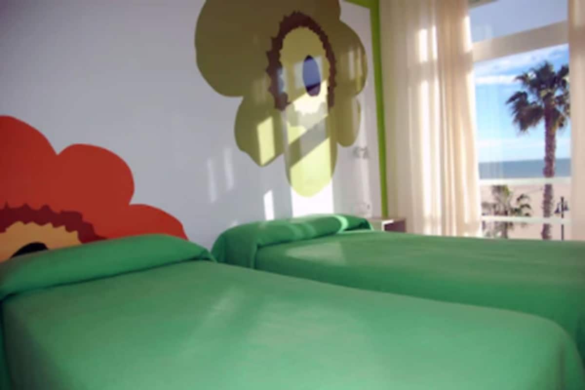 A room with two green beds, a large flower mural on the wall, and a view of the beach through the window.