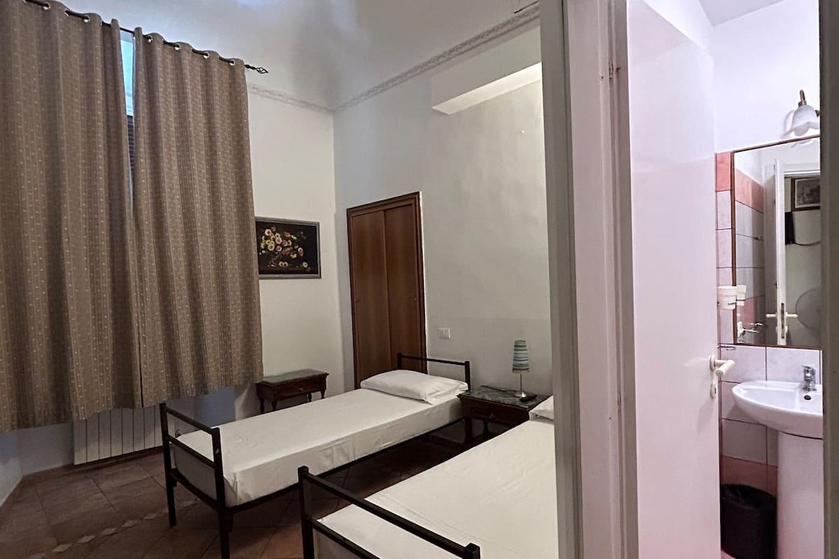 A room with two single beds, a wooden wardrobe, a small table, a lamp, and a bathroom with a sink and mirror.