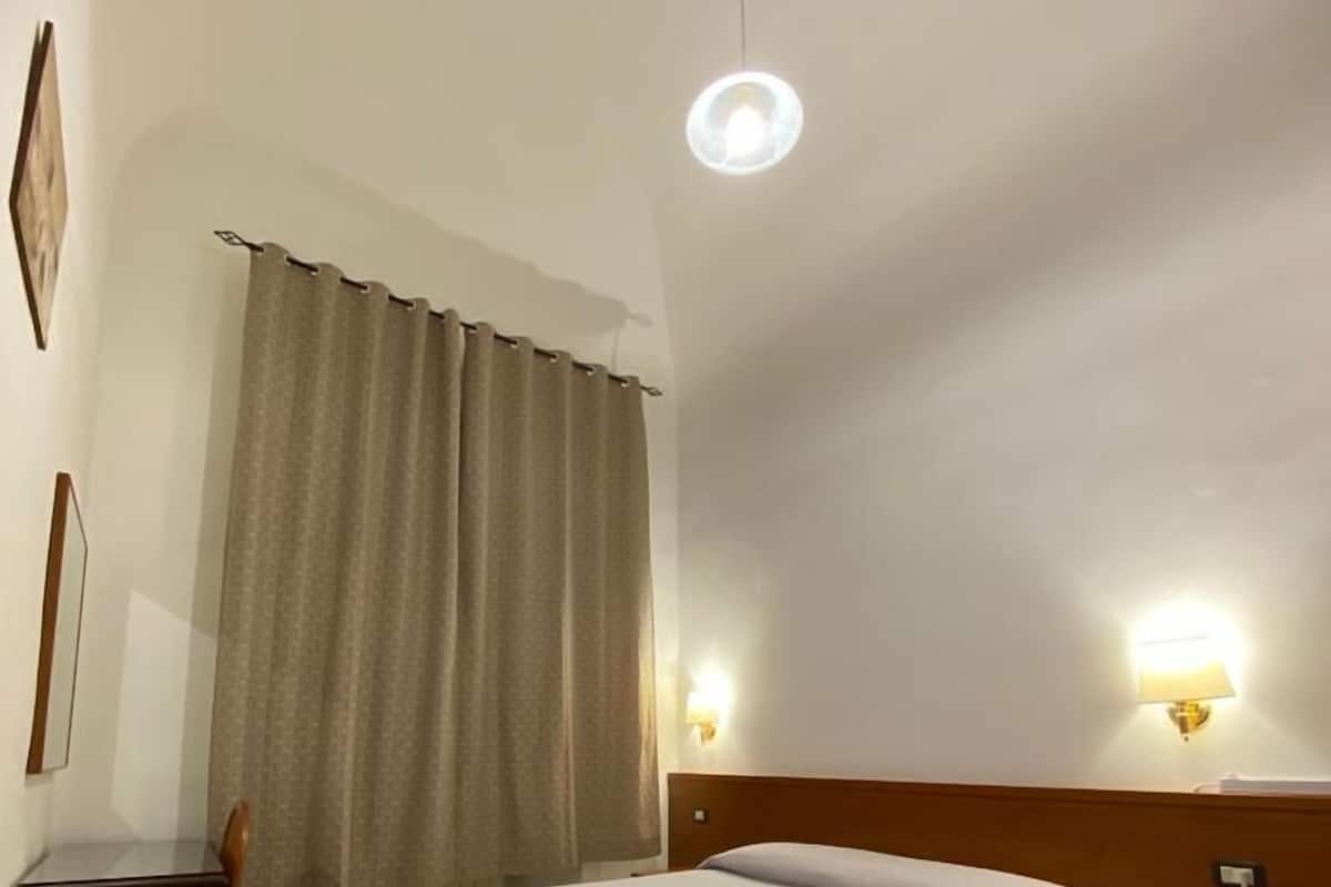 A hotel room with a bed, a desk, a chair, a curtain, and two wall-mounted lights.