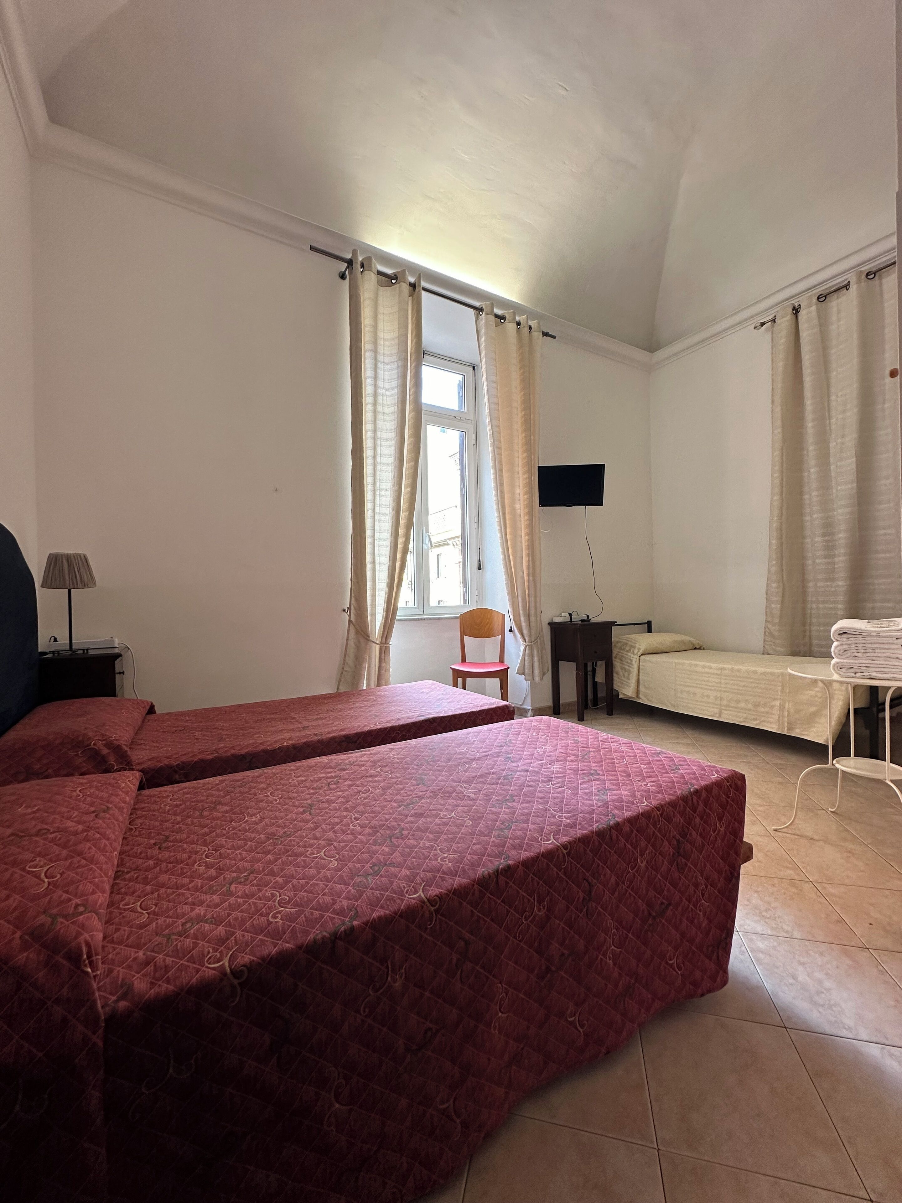 Une chambre avec deux lits, une chaise, une petite table, un téléviseur et une fenêtre avec des rideaux.
