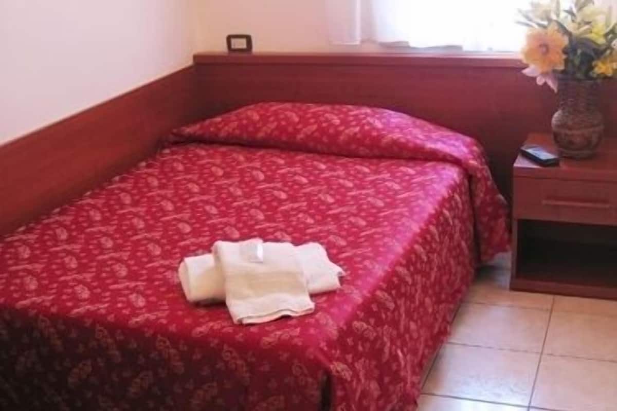 A single bed with a red floral bedspread, a bedside table with a lamp, and a vase with flowers on the nightstand.
