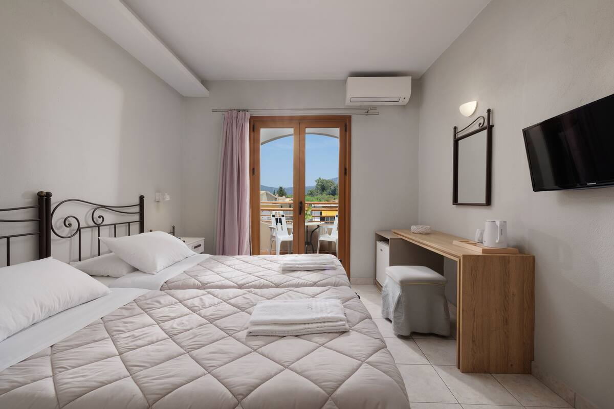 Habitación de hotel con dos camas, un escritorio, una silla, un televisor y vistas a través de una puerta.