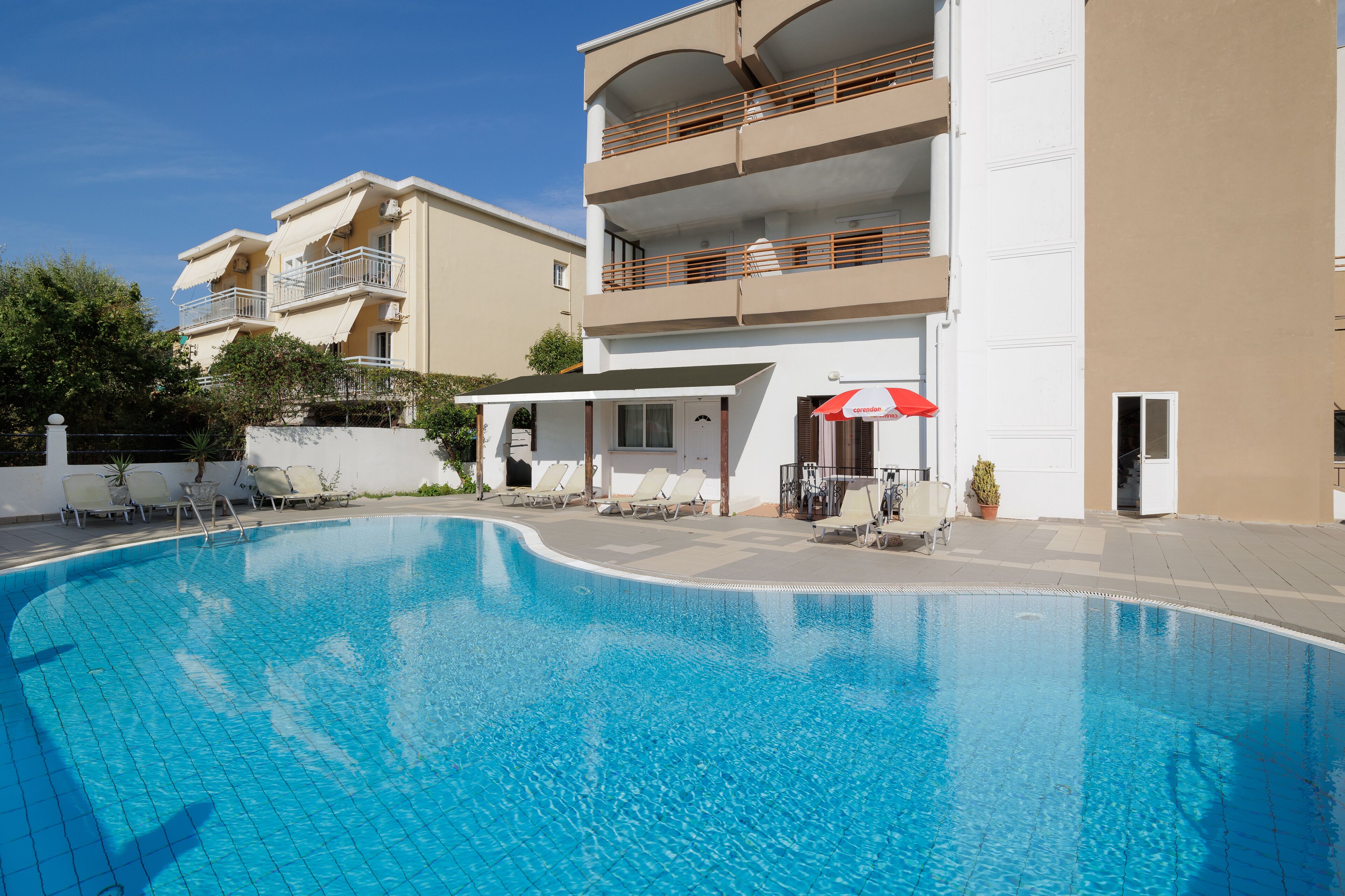 A swimming pool with lounge chairs and umbrellas, surrounded by a building with balconies and a covered area.