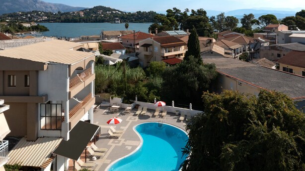 A swimming pool with lounge chairs and umbrellas, surrounded by buildings with red-tiled roofs and a view of a lake and mountains in the background. - Louvre Hotel (Corfu)