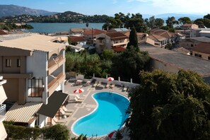 A swimming pool with lounge chairs and umbrellas, surrounded by buildings with red-tiled roofs and a view of a lake and mountains in the background. - Louvre Hotel (Corfu)