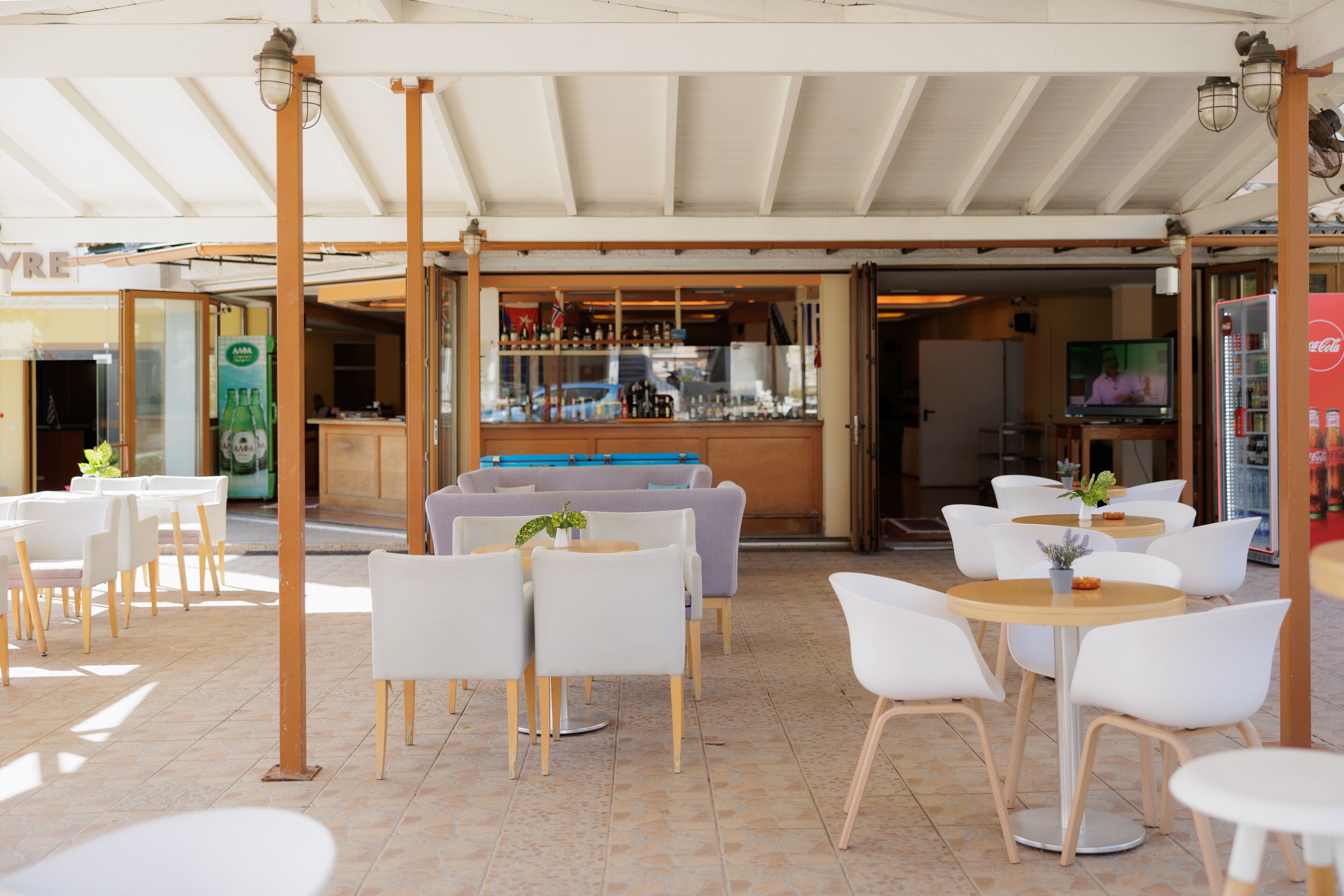 An outdoor cafe with white chairs and tables, a bar area with shelves, and a television screen.