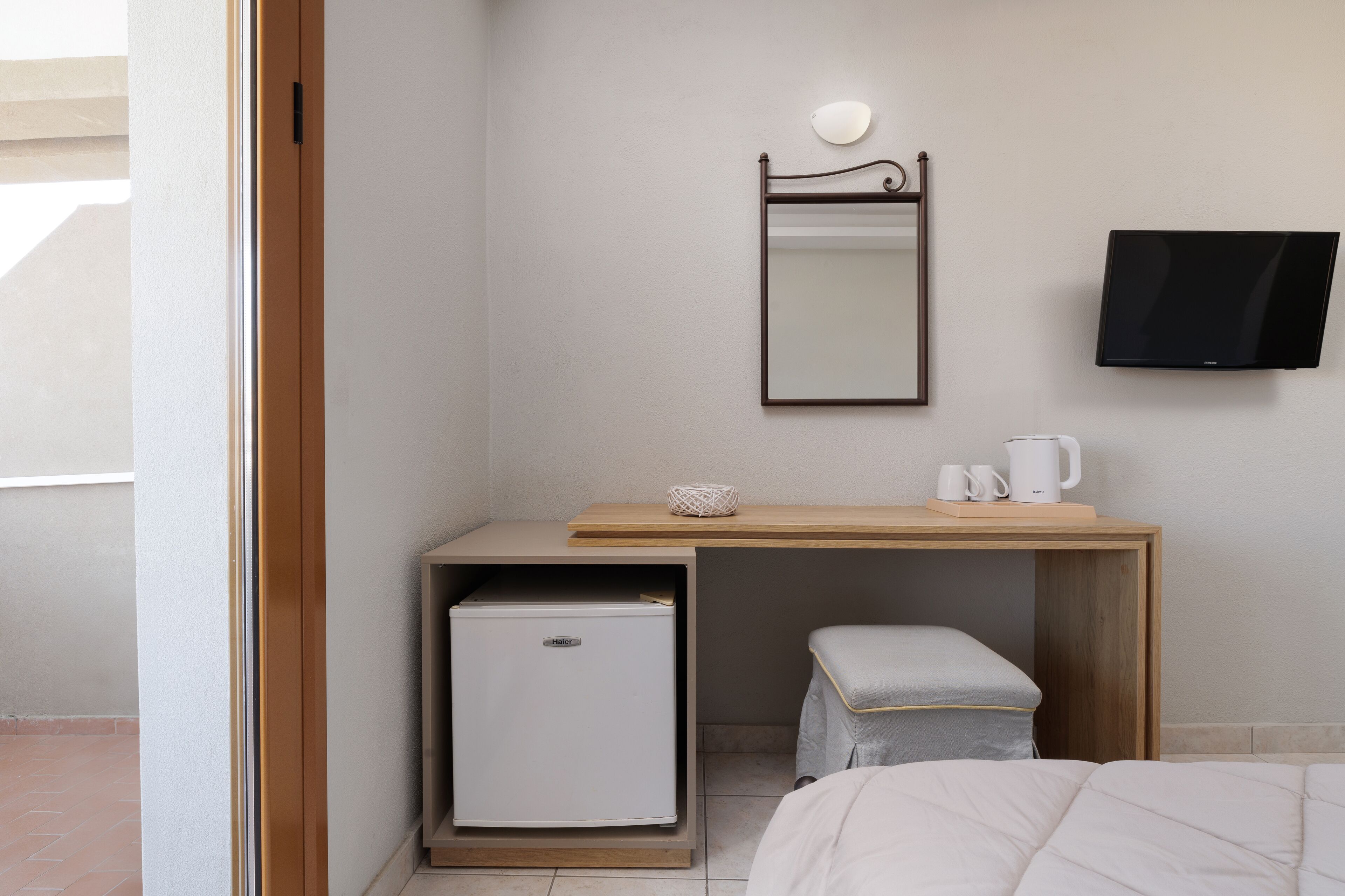 Habitación de hotel pequeña y funcional con cama, una pequeña mesa de trabajo con cafetera, un refrigerador compacto, espejo y televisión en la pared.