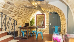 Une pièce aux murs de pierre, avec un escalier en bois, une table entourée de chaises et une porte dotée d’un miroir.