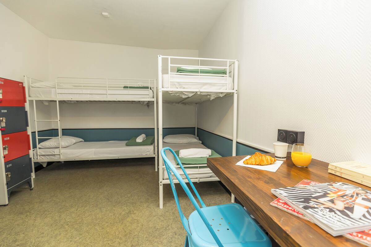 Een studentenkamer met stapelbedden, een bureau met stoel, een tafeltje met een croissant en sinaasappelsap, en een klein boekenrek.