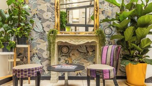 Une pièce avec un papier peint à motifs, une cheminée surmontée d’un miroir, deux fauteuils, une petite table et plusieurs plantes en pot.
