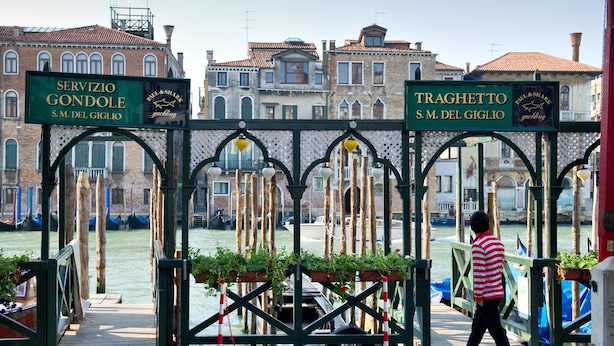 A pedestrian bridge with signs for "Servizio Gondole" and "Traghetto S.M. del Giglio" over a canal, with buildings in the background. - Palazzo del Giglio (Venice)