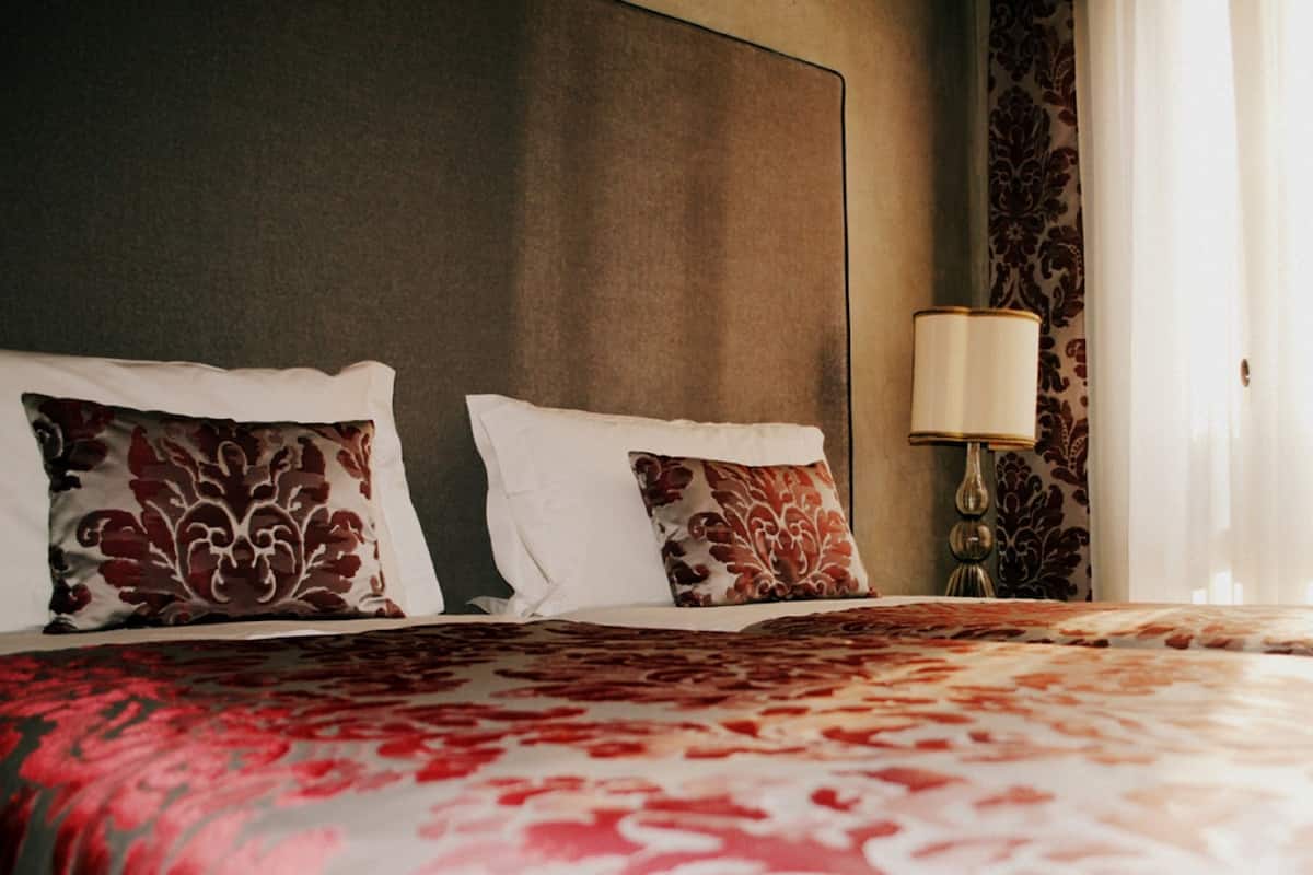 Une chambre d’hôtel avec un grand lit, deux oreillers à motifs floraux, une lampe de chevet et une fenêtre avec des rideaux.