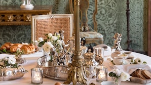 A table set with fine china, silverware, and candles, with a painting and lamps in the background. - Residenza Napoleone III (Rome)