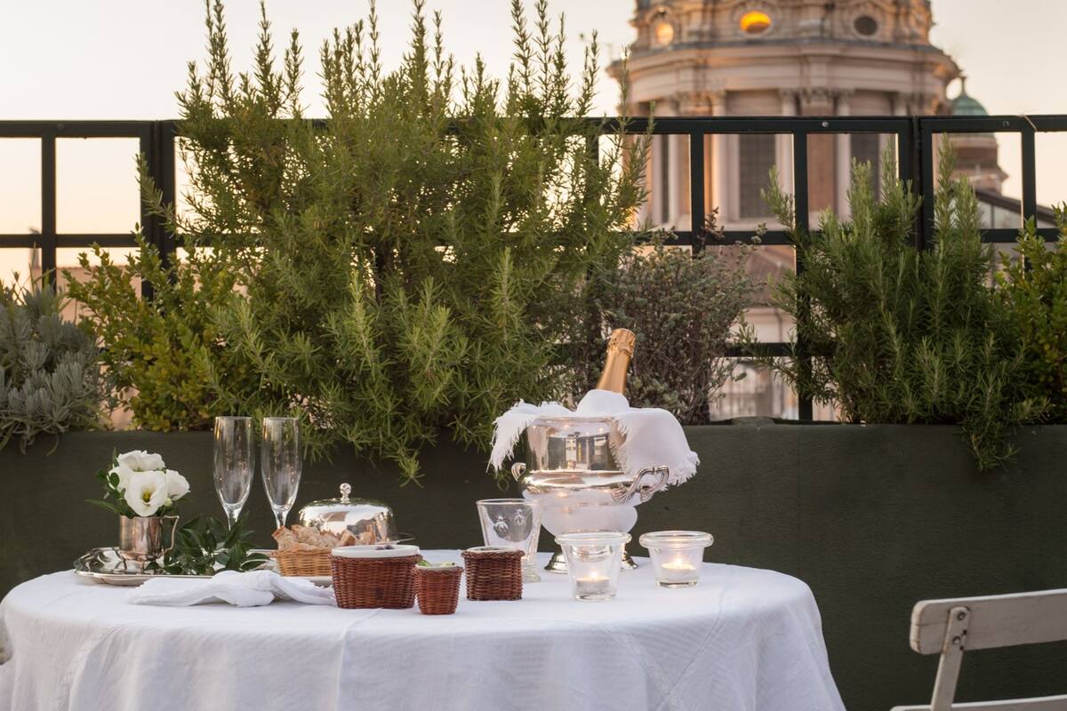 An outdoor dining setup with a table set for two, featuring a bottle of champagne, glasses, and a basket of bread. A large dome structure is visible in the background.