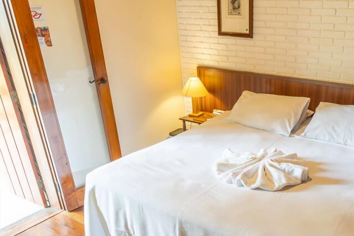 A neatly made bed with white linens and a towel, a wooden headboard, a nightstand with a lamp, and a brick-patterned wall with framed pictures.