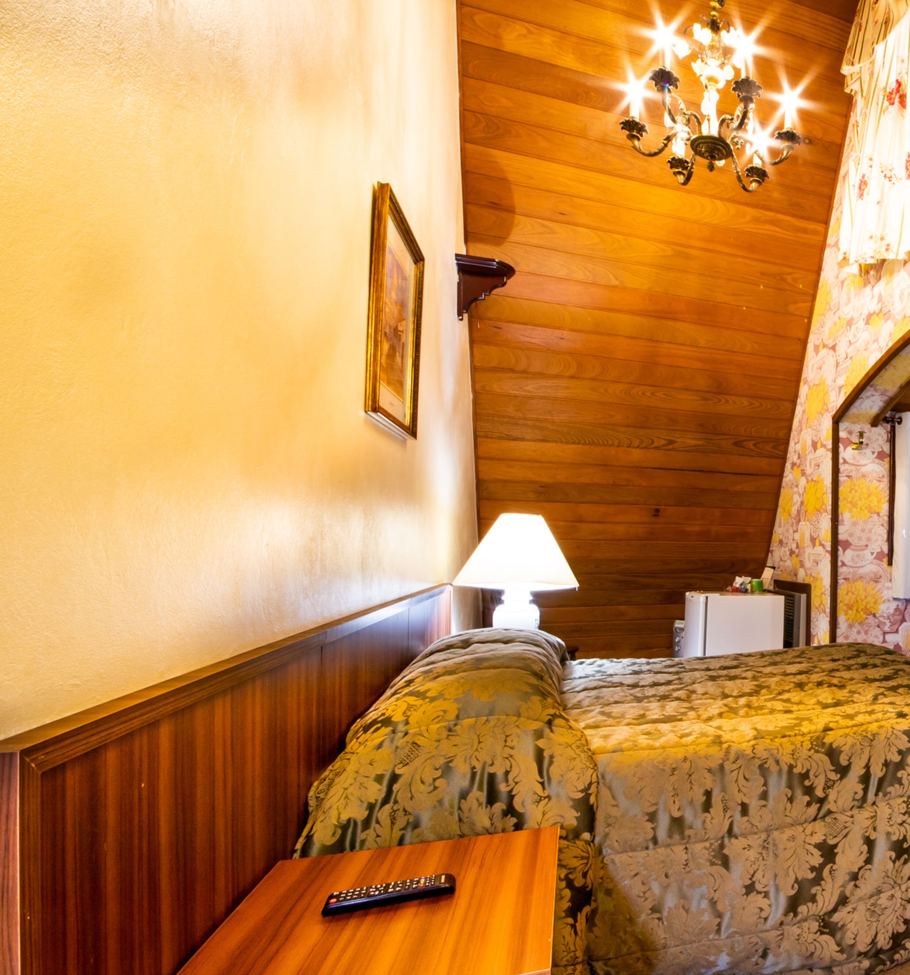 Um quarto aconchegante com teto de madeira, cama com roupa de cama florida, mesa de cabeceira com abajur e uma moldura na parede.