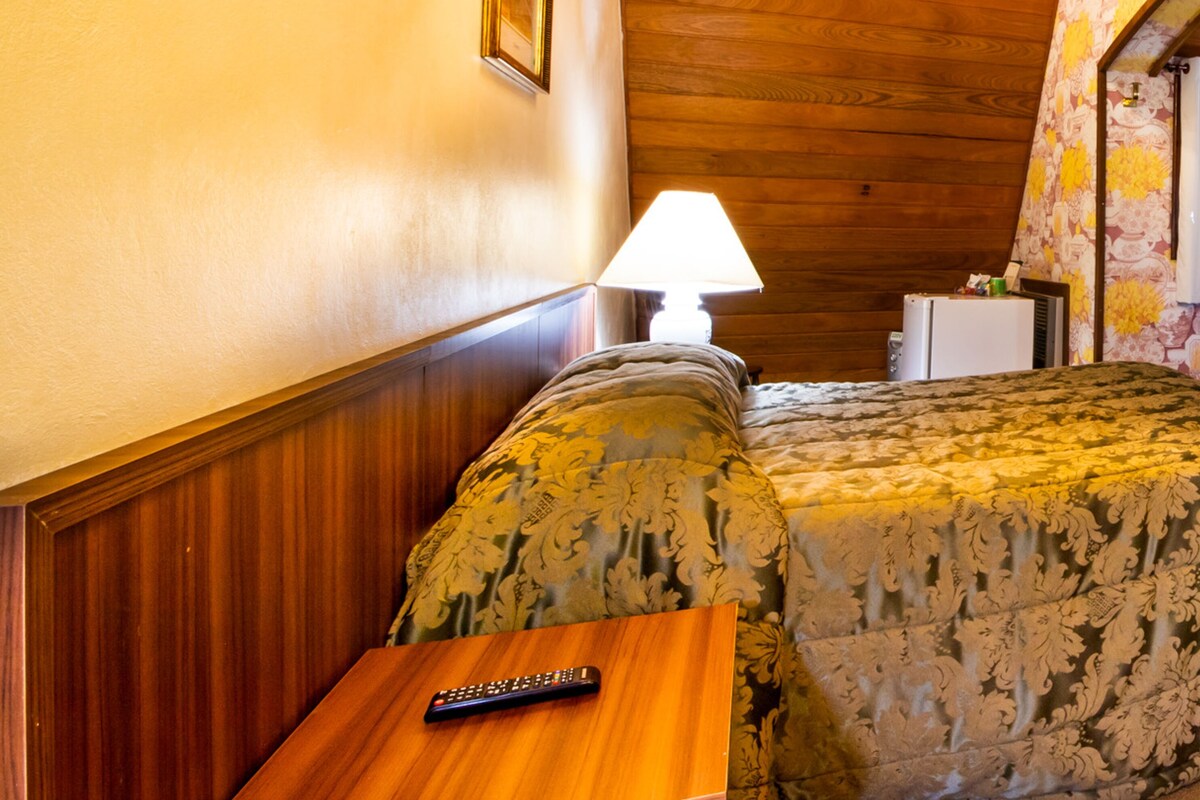 Una habitación acogedora con techo de madera, una cama con ropa de cama floral, una mesita de noche con lámpara y un cuadro enmarcado en la pared.