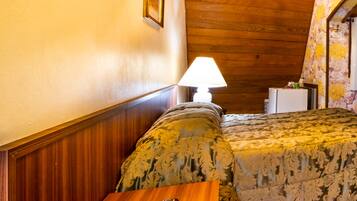 Um quarto aconchegante com teto de madeira, cama com roupa de cama florida, mesa de cabeceira com abajur e uma moldura na parede.
