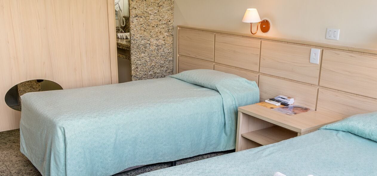 Een hotelkamer met twee enkele bedden, een houten hoofdbord, een nachtkastje met een lamp en een klein tafeltje met een tijdschrift.