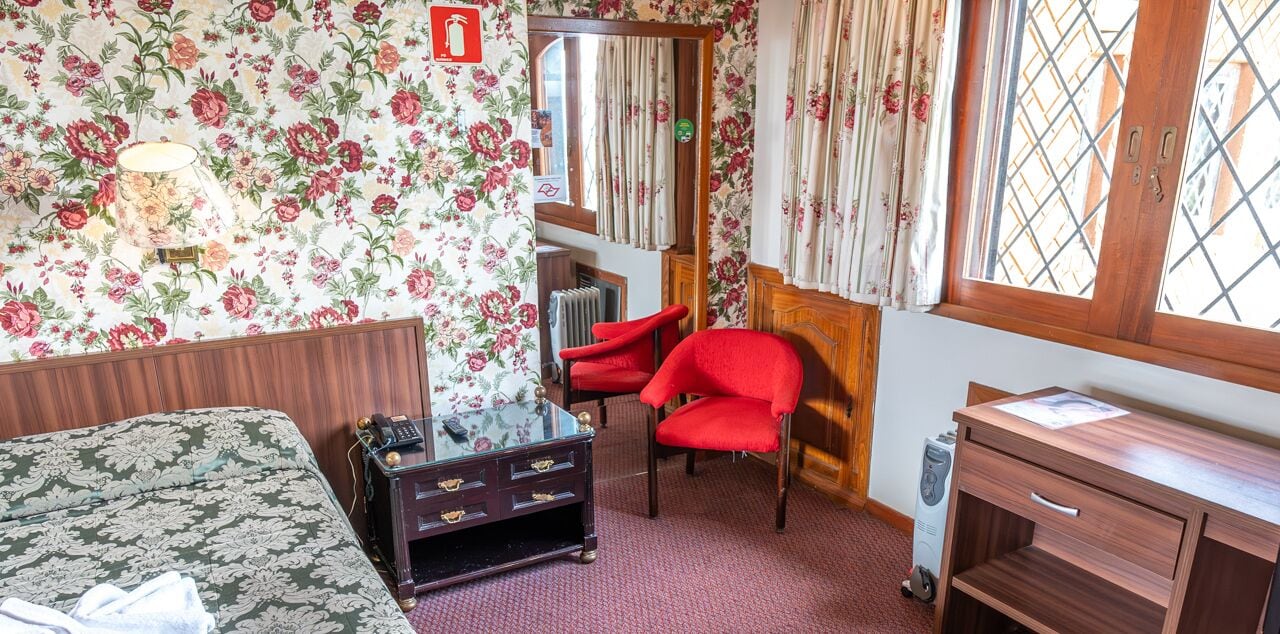A room with floral wallpaper, a bed, a red chair, a wooden dresser, and a radiator.
