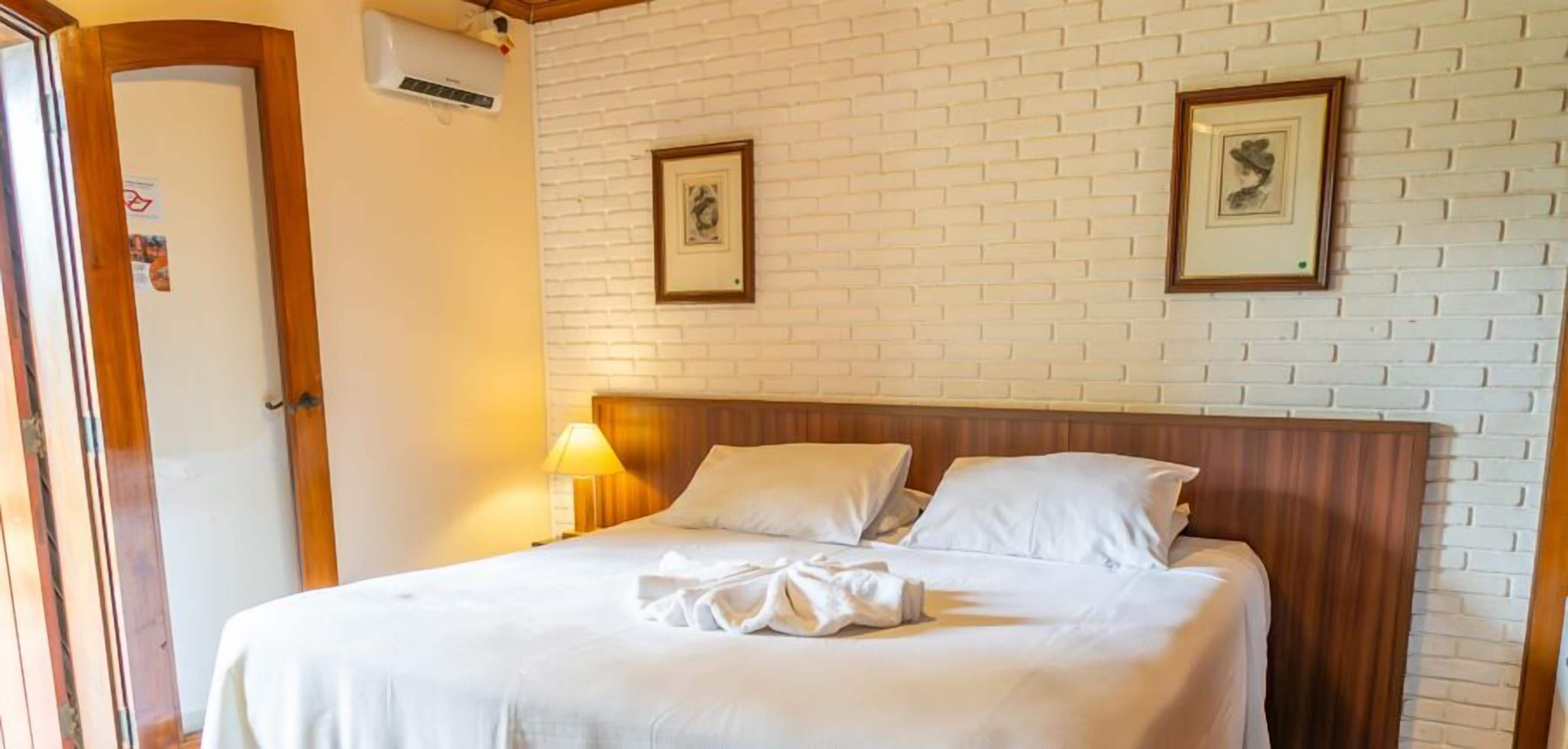 A hotel room with a white brick wall, a wooden headboard, two framed pictures, a bed with white linens, and a lamp on the nightstand.