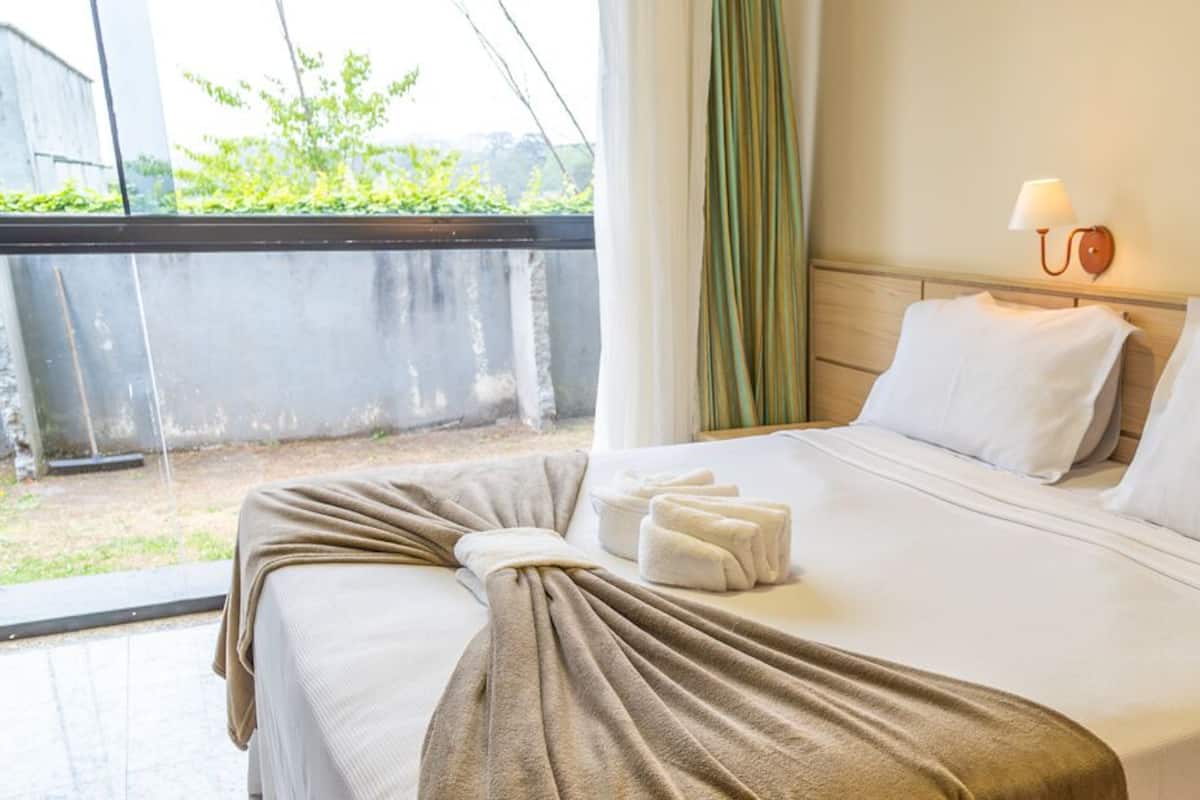 A neatly made bed with white linens and a brown throw blanket, a bedside lamp, and a view of greenery outside.