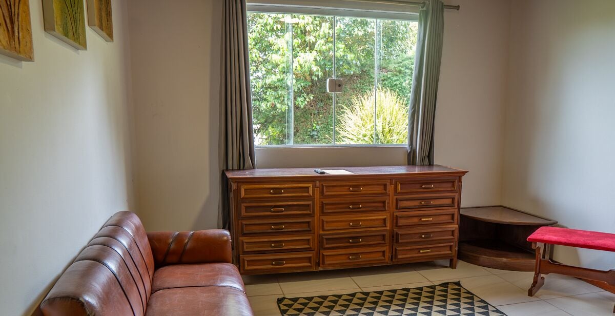 A room with a brown leather sofa, a wooden dresser, a red bench, and a window with a view of greenery.