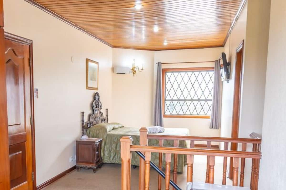 A bedroom with a wooden ceiling, a bed with a green bedspread, a nightstand, and a window with a metal grid.