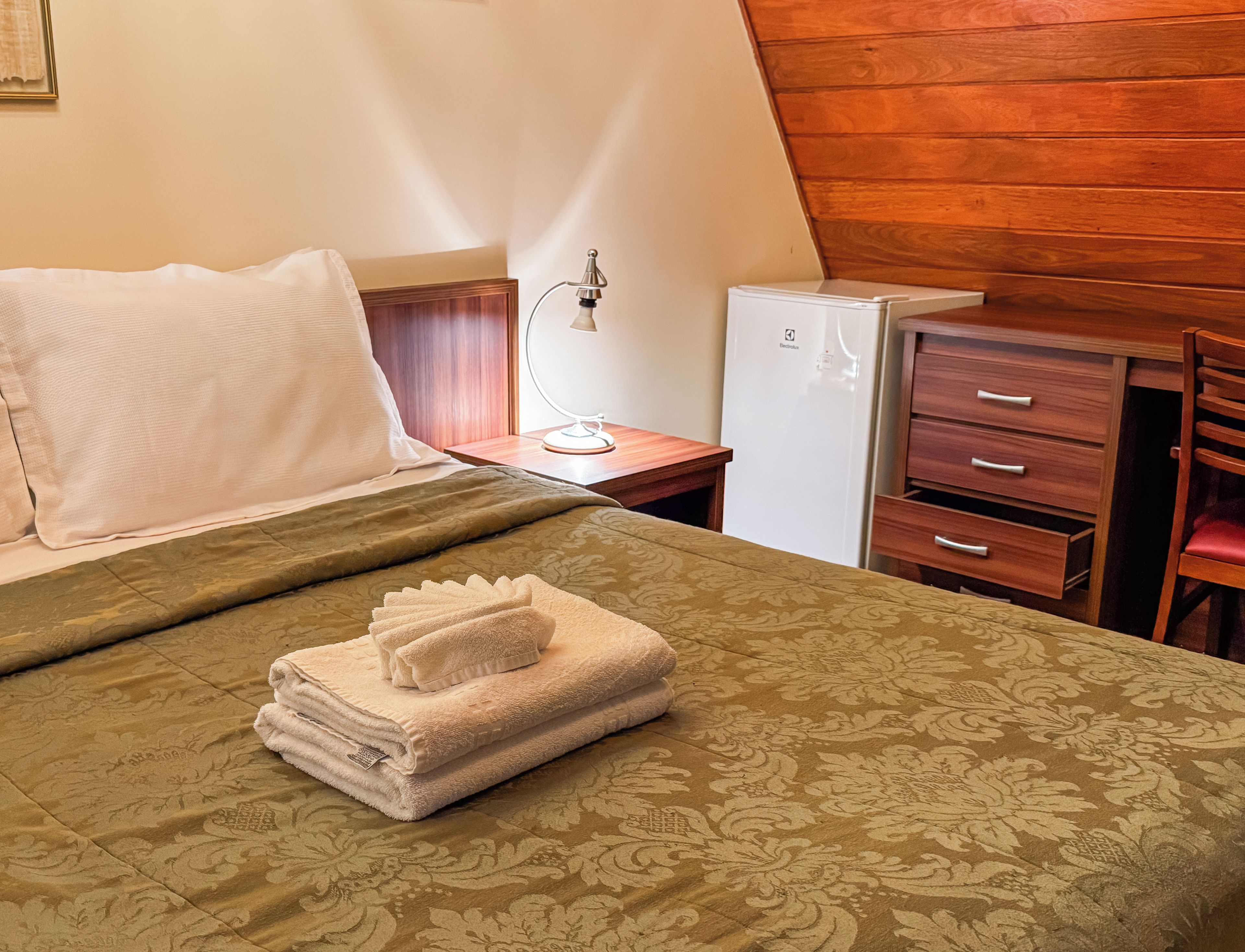 A hotel room with a bed, a desk, a lamp, a refrigerator, and a chair.