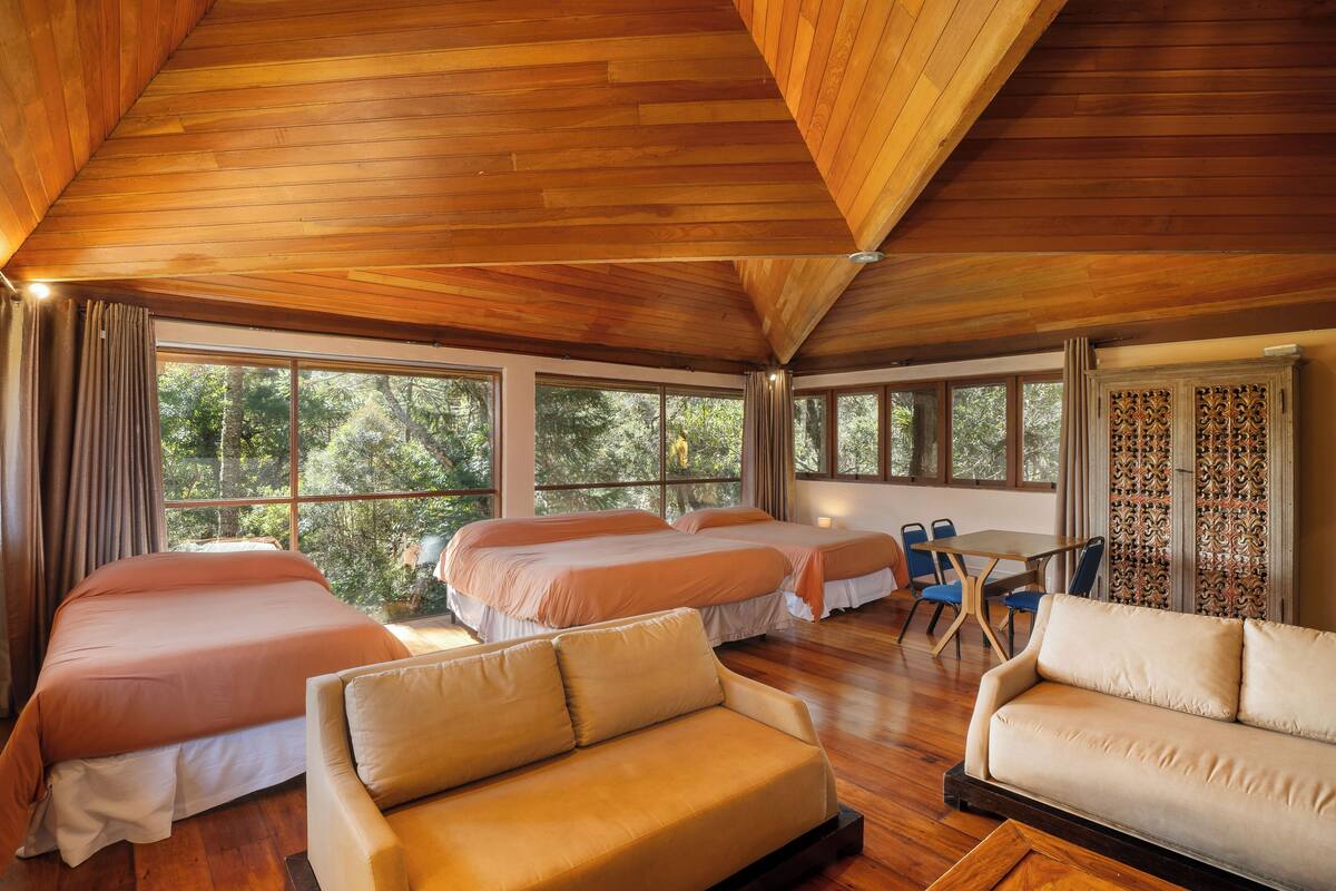A spacious room with wooden ceilings, two beds, a sofa, and a dining area with a view of trees.