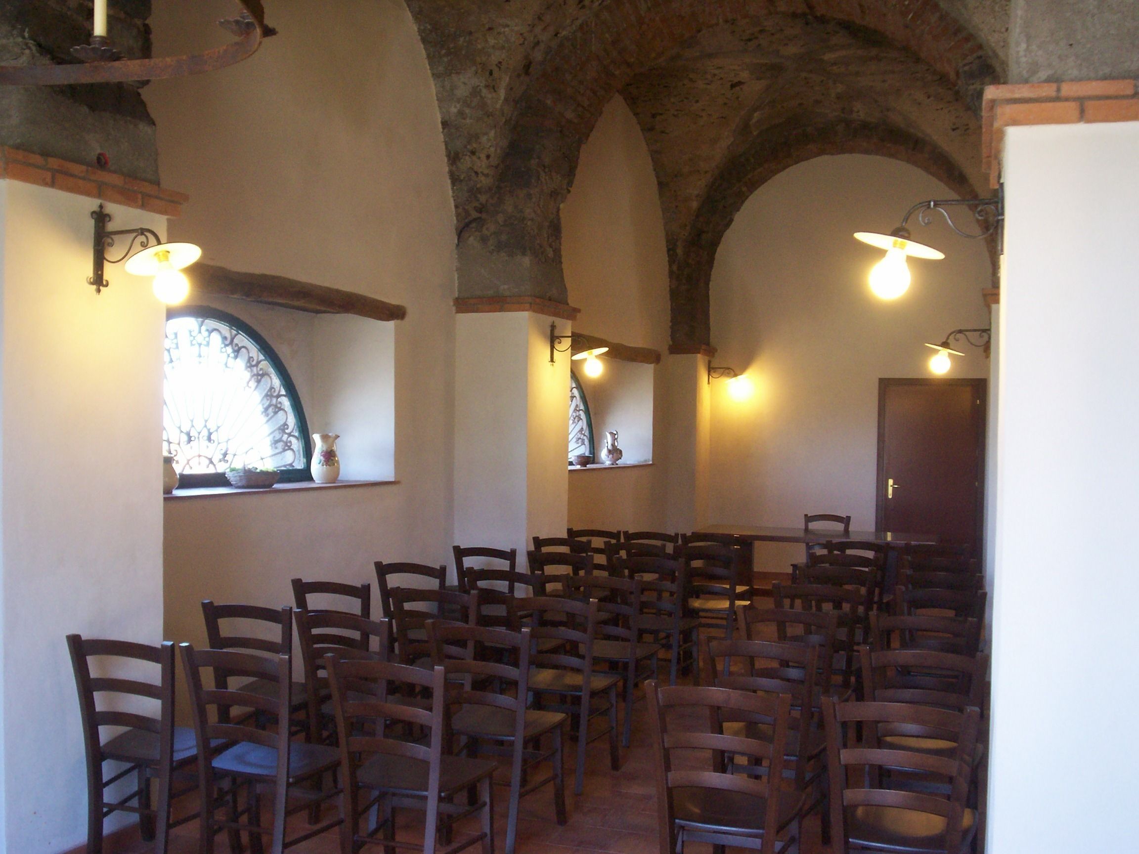Une pièce avec des rangées de chaises en bois disposées pour accueillir les personnes, une arche en pierre et des luminaires fixés au mur.