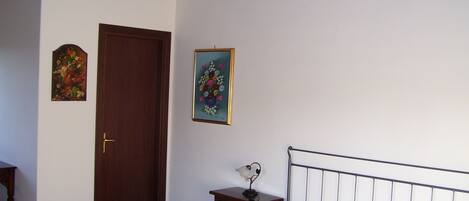 A bedroom with a bed, a wooden door, a small bedside table with a lamp, and two framed floral paintings on the wall.