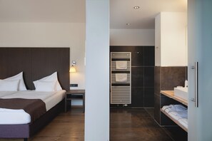A modern hotel room with a large bed, bedside table, and a built-in bench. There is a separate bathroom with a shower, sink, and towel rack. - GolfHotel Gut Neuenhof (Fröndenberg/Ruhr)