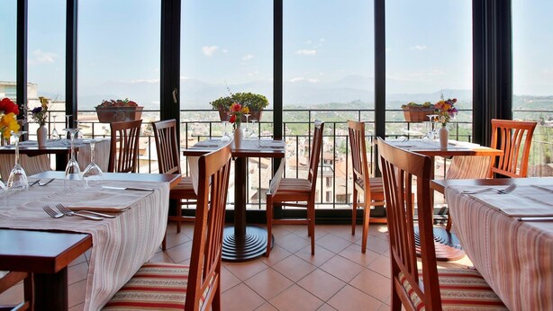 A restaurant with large windows offering a scenic view. Tables are set with chairs and potted plants. - Hotel Garibaldi (Frosinone)