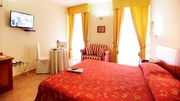 Une chambre d’hôtel avec un grand lit, une petite table sur laquelle est posé un vase de fleurs, une chaise, un téléviseur fixé au mur et des rideaux.