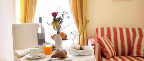 Una mesa con desayuno, una botella, un florero con flores y un sofá a rayas al fondo.