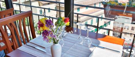 Una mesa de comedor dispuesta con un jarrón de flores, copas de vino y cubiertos en un balcón con vistas a la ciudad.