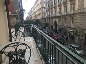 A balcony with a metal railing overlooking a street with parked cars and pedestrians.