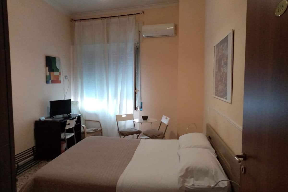 A hotel room with a bed, a desk, a chair, a television, and an air conditioning unit.