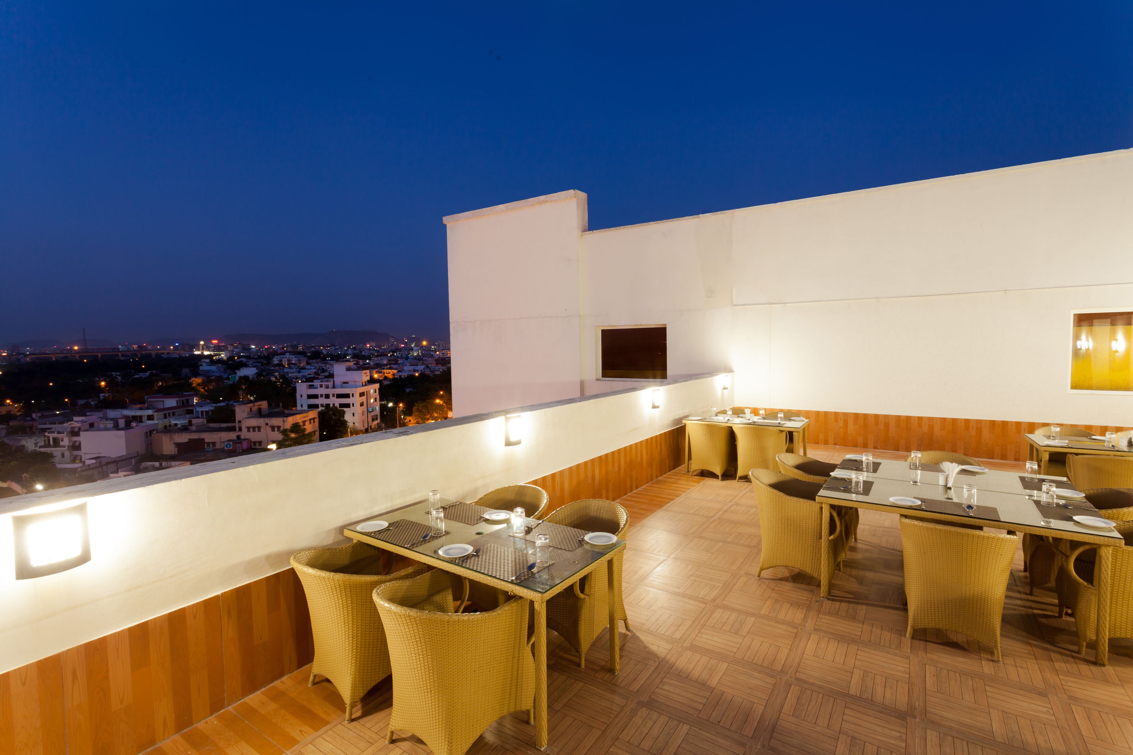 A rooftop terrace with tables and chairs, a city view, and a wooden floor.