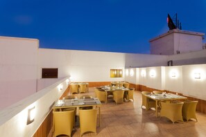 Ein Dachterrasse-Essbereich mit mehreren Tischen und Stühlen, beleuchtet durch an Wänden befestigte Lampen.