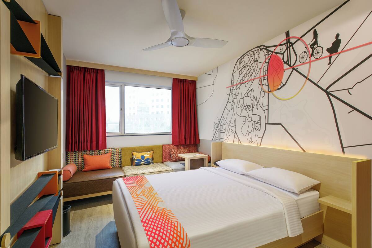 Habitación de hotel con cama, televisión, escritorio, silla, sofá y ventana con cortinas.