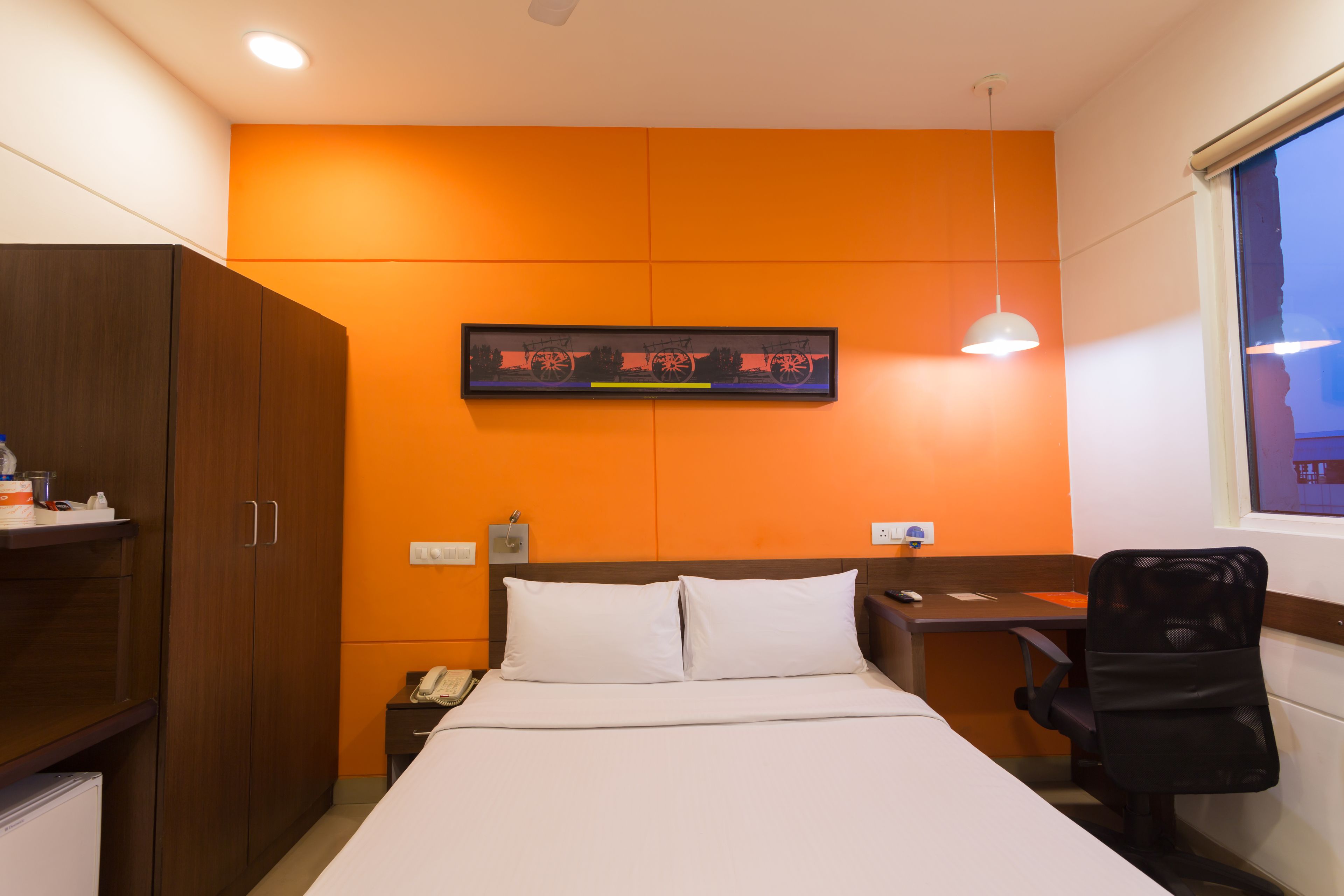A hotel room with a bed, a desk, a chair, a wardrobe, a painting on the wall, and a window.