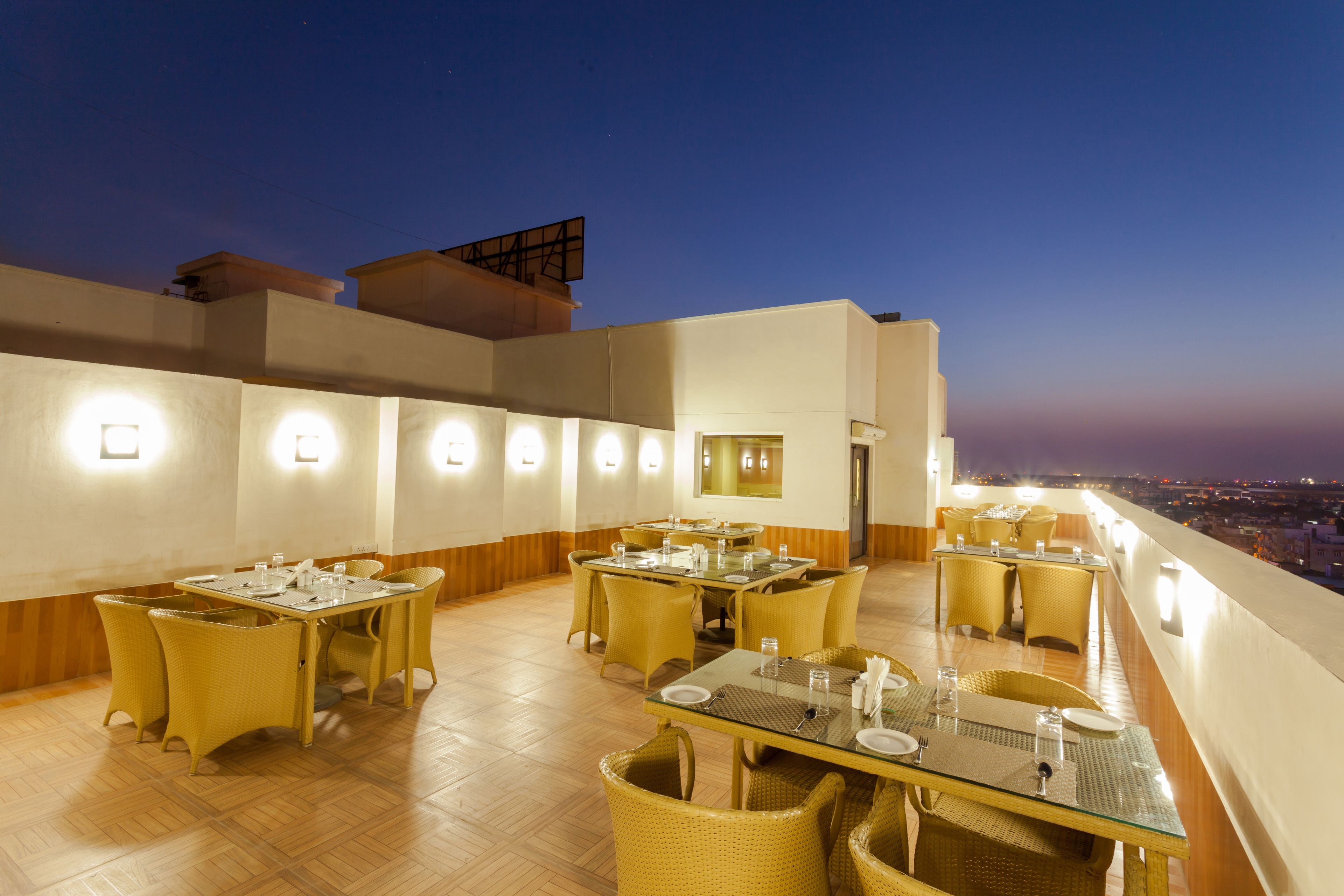 Uma área de refeições no terraço com várias mesas e cadeiras, iluminada por luminárias fixadas na parede, com vista para a paisagem urbana ao entardecer.
