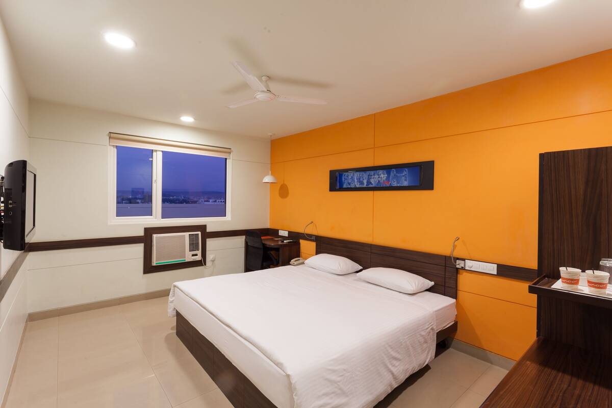 Habitación de hotel con cama, escritorio, silla, ventana con vistas, ventilador de techo y televisor montado en la pared.