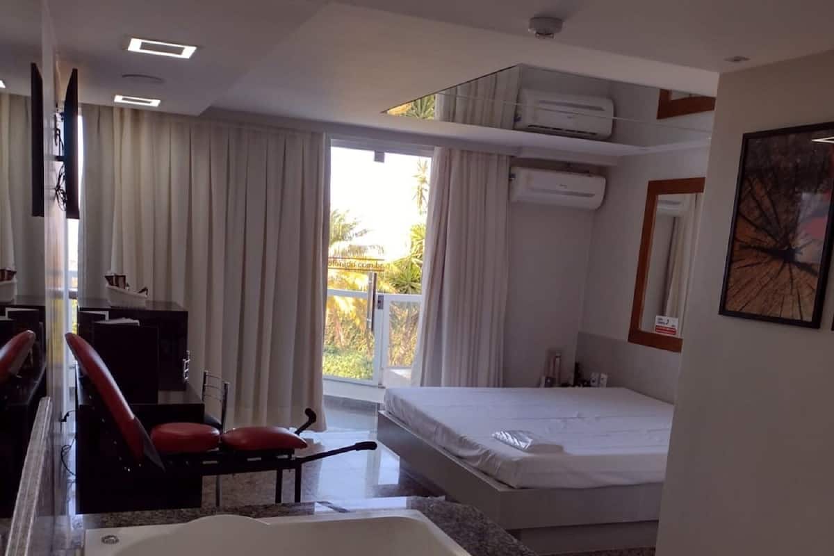 A hotel room with a large bathtub, a bed, a desk with a chair, and a view of the outdoors through a sliding door.